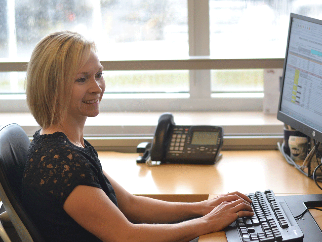 A woman smiling, is typing on a keyboard with a telephone in the background and half of the desktop is visible