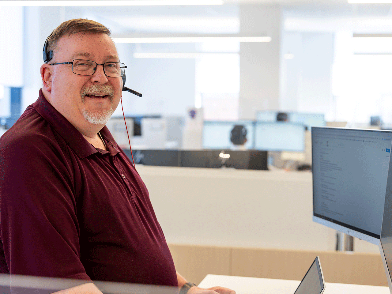 Smiling man wearing a headset in a modern office, standing at a desk with a laptop and monitor.