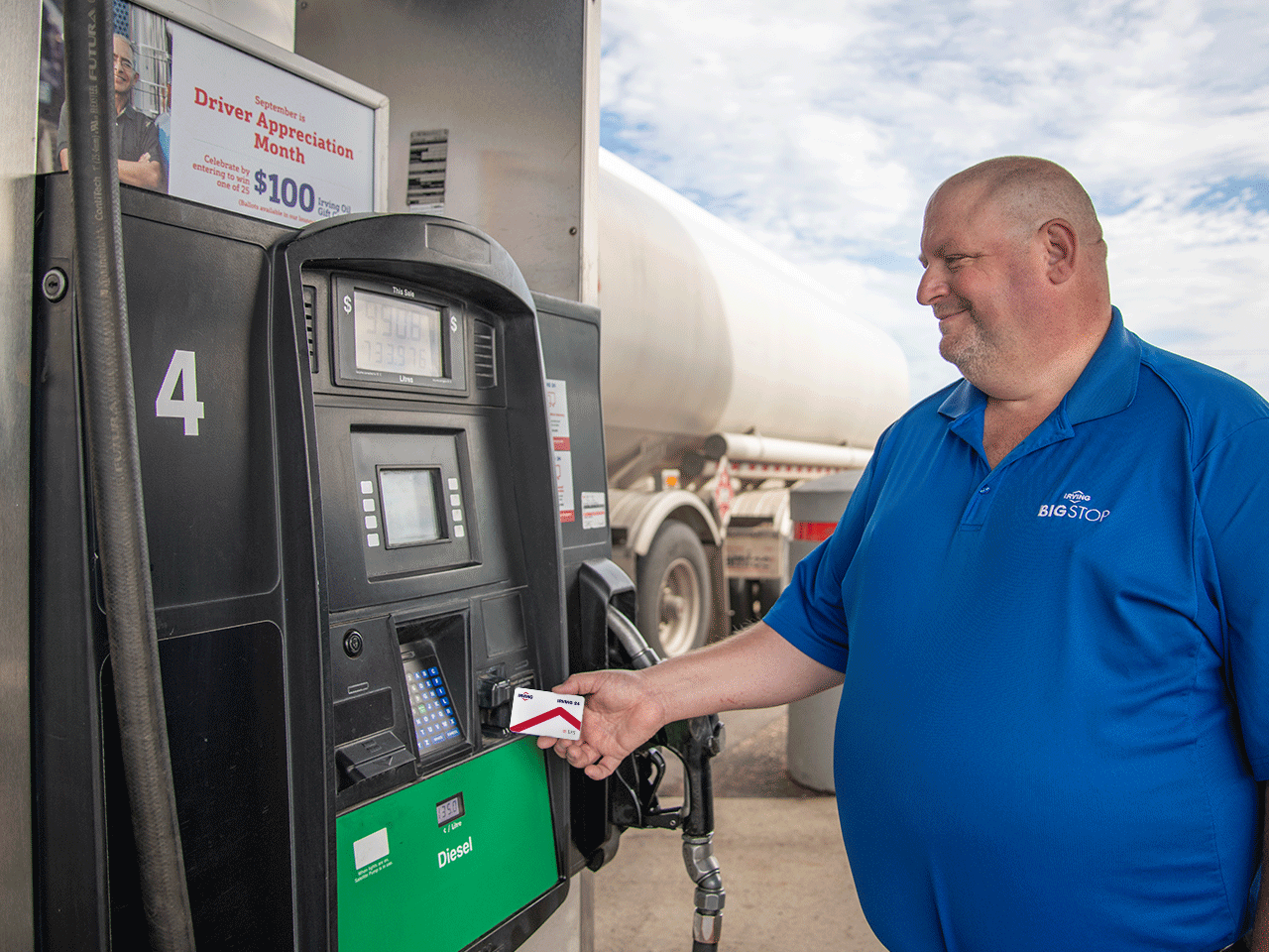 A customer at an Irving 24 station is using Irving 24 card, he is happy and wearing a blue color t-shirt. A truck is parked behind him under a blue cloudy sky.
