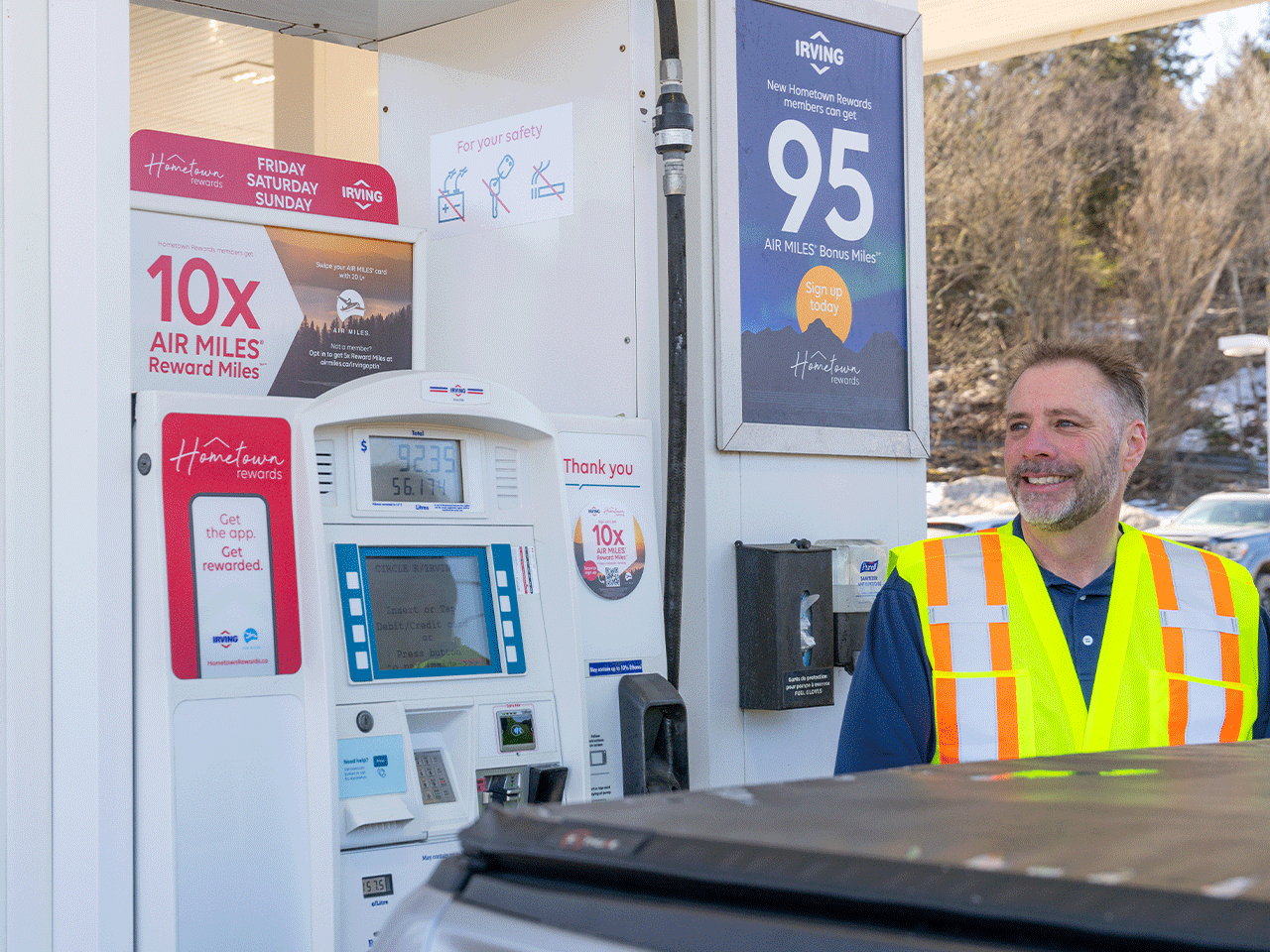 A man standing at the gas pump, looking away from the camera wearing a safety vest and the half trunk of a SUV is in the foreground and there are various Ads on the gas pump behind that man. The trees are in the far background of o n the 