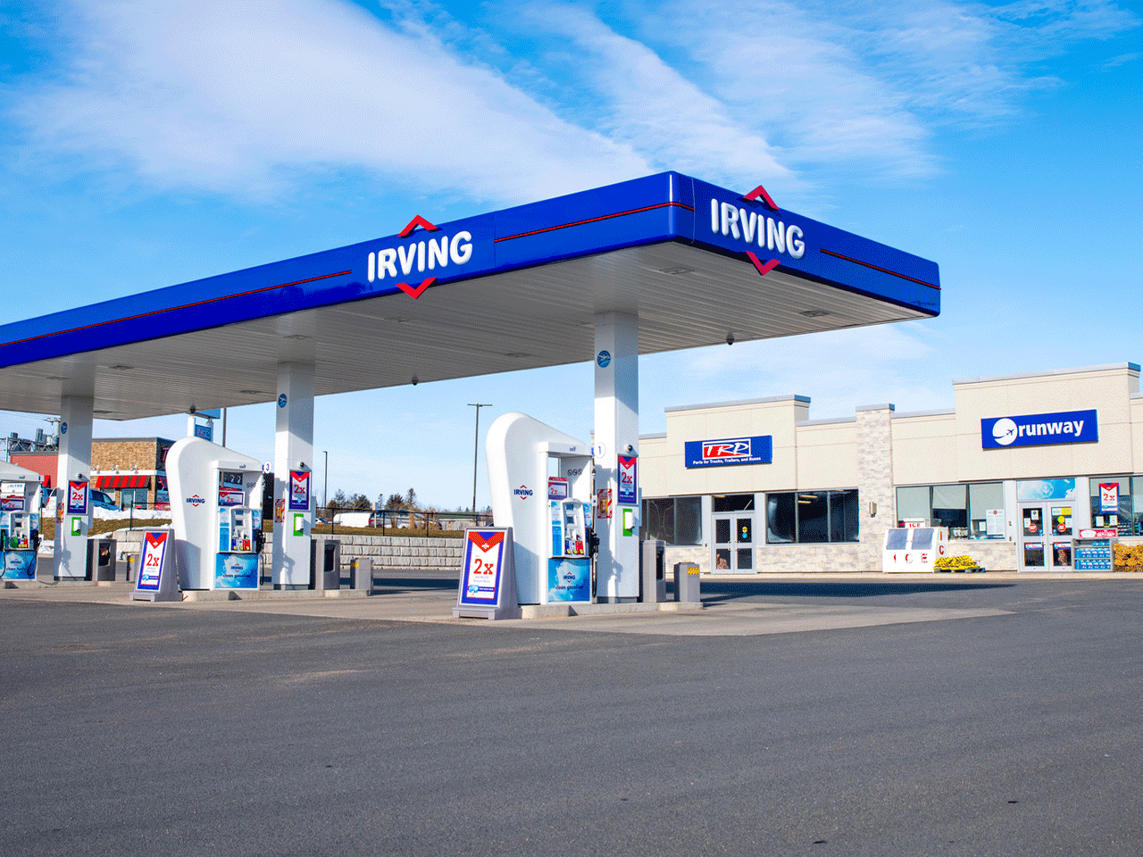 Wide-angle of Irving gas station with blue sky and few scattered clouds and a road in the foreground with a convenient store behind the gas pump. 