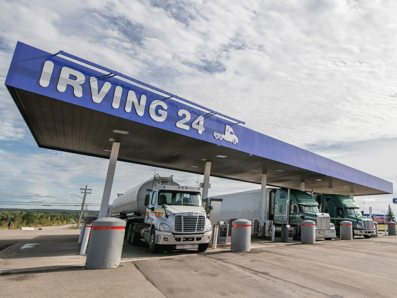 Three trucks parked in an Irving 24 forecourt fuel pump on a cloudy sunny day. 