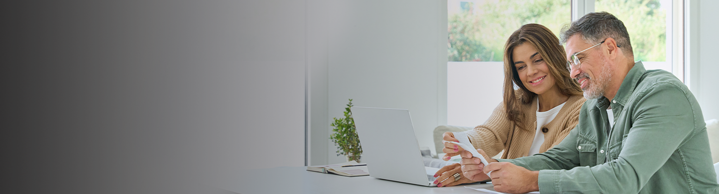 Two people sit at a table using a laptop and reviewing a credit card; a small plant and notebook sit nearby, with soft daylight from a window in the background.
