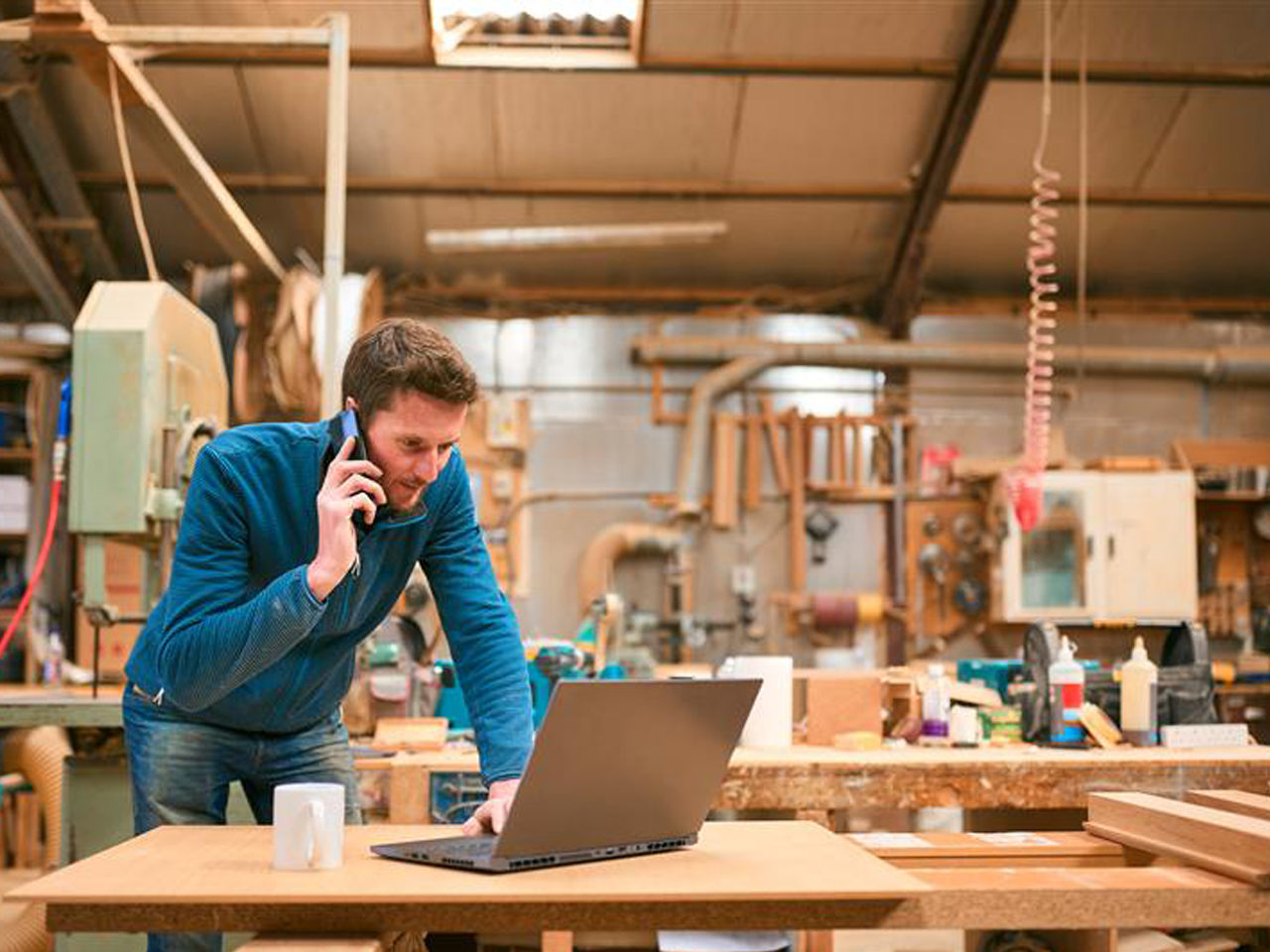 Man in a workshop talking on the phone while working on a laptop, surrounded by woodworking tools and machinery.