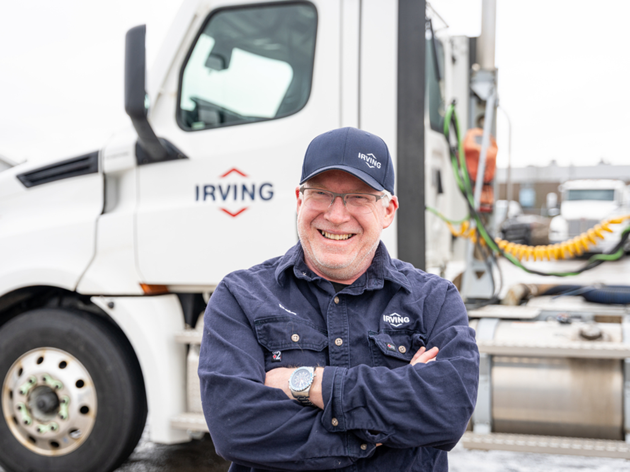 Person in an Irving work uniform standing with arms crossed in front of an Irving transport truck, with the truck cab and equipment visible in the background.